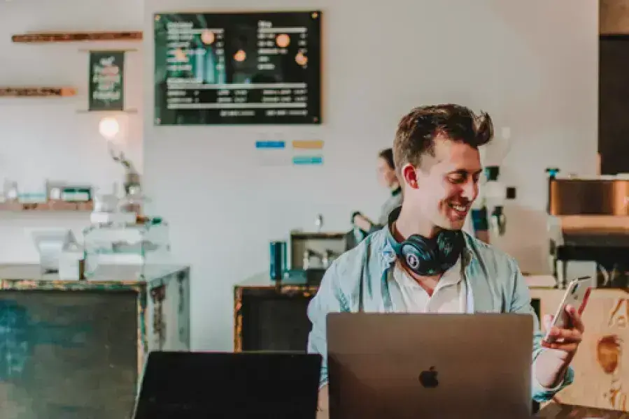 A person on their laptop smiling while looking at their cell phone